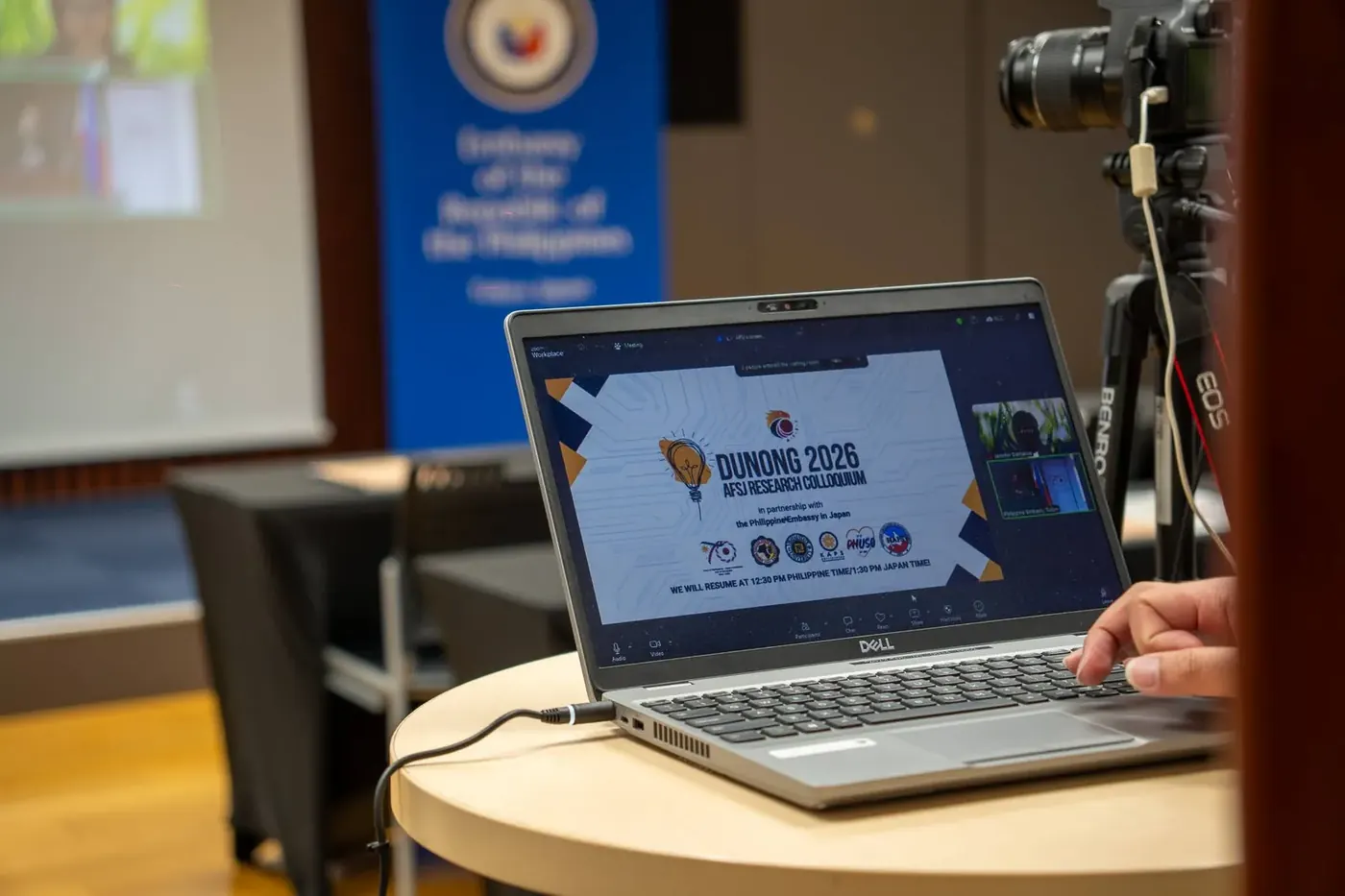 A laptop at the Philippine Embassy in Tokyo showing the DUNONG 2026 AFSJ Research Colloquium opening slide, with a camera on a tripod beside it